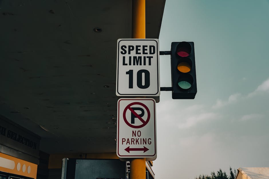 A vertical yellow pole supporting a traffic light with three coloured sections—red, yellow, and green—mounted on the corner of a building under an overhang. Attached to the pole are two rectangular signs: the upper one displays a white background with black text indicating a speed limit of 10 miles per hour, and the lower sign features a white background with a red circle and a slash through the letter 'P,' accompanied by the message 'No Parking' in red text and left and right arrows indicating no parking zones. The scene is set outdoors during daytime with a partially cloudy sky visible in the background; the environment suggests an urban or suburban location suitable for house removals or furniture transport, consistent with the services of Man With a Van West Ham.