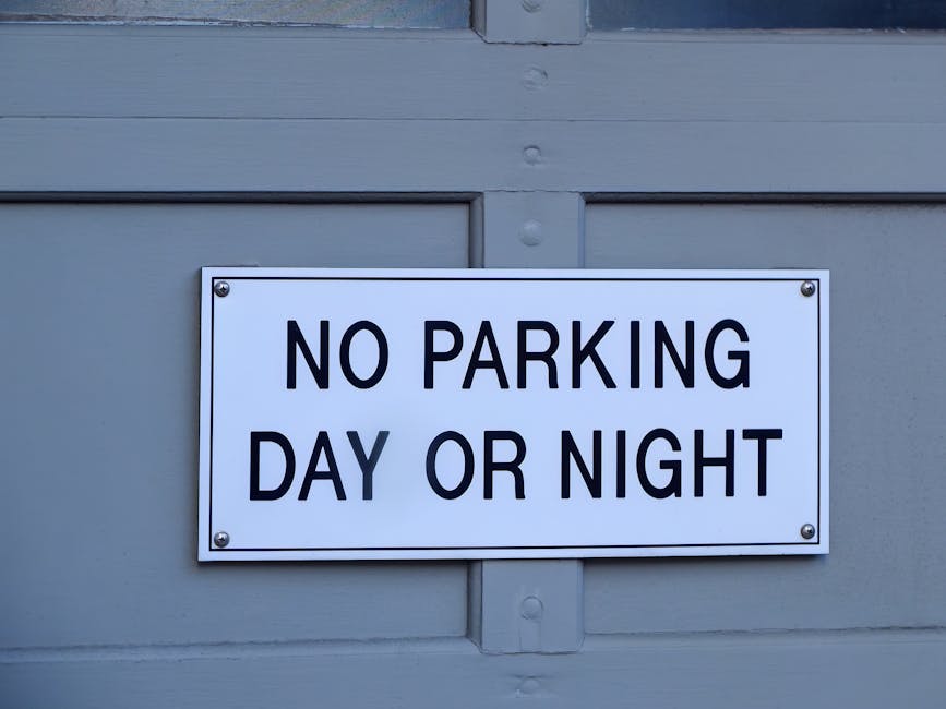 A close-up view of a rectangular white metal parking restriction sign mounted on a grey wooden wall, displaying the message 'NO PARKING DAY OR NIGHT' in bold black capital letters. The sign is secured with four screws, one at each corner. The background features horizontal wooden panels painted grey, typical of a garage or building exterior. The lighting is natural, indicating it is daytime. This setting suggests a location where the property owner or business, such as Man With a Van West Ham, might conduct house removals or furniture transport, emphasizing the importance of adhering to parking regulations when carrying out home relocation or moving services in the West Ham area.