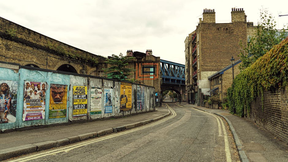 A narrow residential street with a gently curved asphalt surface, featuring double yellow lines along the curbside. On the left, a long weathered metal fence covered in colorful posters and advertisements separates the sidewalk from an industrial brick building with arched windows. On the right, a brick wall partially covered with green ivy runs along a residential property, with a small tree extending over the pavement. In the background, there is a blue metal bridge spanning above the street, connecting two brick buildings with multiple stories and chimney stacks. The scene is well-lit with natural daylight, typical of an overcast sky, providing a clear view of urban environments suitable for house removals, furniture transport, and home relocation planning, as often managed by companies like Man With a Van West Ham.