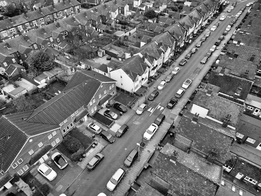 An aerial black-and-white photograph of a residential street showing a row of terraced houses with pitched roofs and small front gardens. Parked cars line both sides of the narrow street, with some vehicles parked on the pavement itself. The road features a central double white line and a pedestrian crossing near the bottom of the image. The houses and streets are tightly packed, with some greenery visible in the form of small trees and garden shrubs. The scene depicts a typical UK urban residential area, suitable for house removals and furniture transport, as with services from Man With a Van West Ham. The lighting suggests daytime, with clear visibility of the entire street, which is useful for planning home relocation logistics and loading areas during a move.