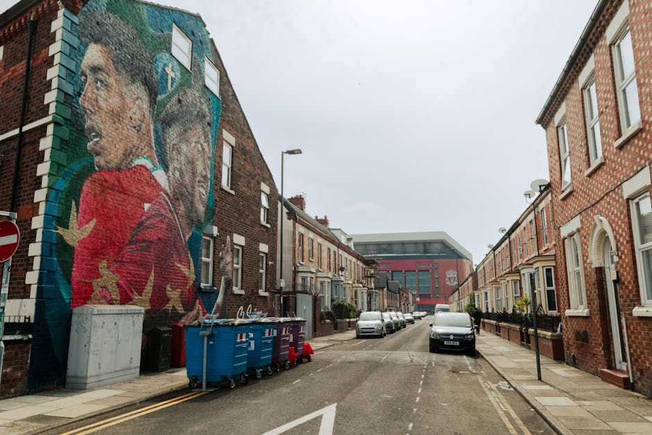 A narrow residential street with a gently curved asphalt surface, featuring double yellow lines along the curbside. On the left, a long weathered metal fence covered in colorful posters and advertisements separates the sidewalk from an industrial brick building with arched windows. On the right, a brick wall partially covered with green ivy runs along a residential property, with a small tree extending over the pavement. In the background, there is a blue metal bridge spanning above the street, connecting two brick buildings with multiple stories and chimney stacks. The scene is well-lit with natural daylight, typical of an overcast sky, providing a clear view of urban environments suitable for house removals, furniture transport, and home relocation planning, as often managed by companies like Man With a Van West Ham.