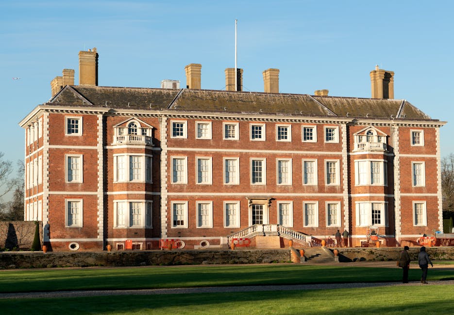 A large, historic brick building with a symmetrical facade, multiple sash windows, and ornate architectural details such as decorative stonework and small balconies. The building is set against a clear blue sky, with a well-maintained lawn and paved pathway in the foreground. The ground level features a central entrance with a staircase leading up to a door, and the surrounding area includes a few people walking nearby. This setting illustrates the type of property involved in residential house removals, with ample space for loading and unloading furniture and boxes, highlighting the suitability of services offered by Man With a Van West Ham for home relocation and furniture transport within local areas.