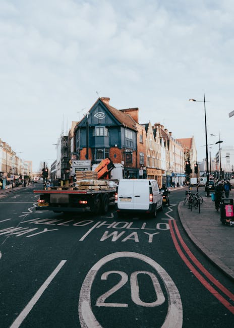 A flatbed removal truck loaded with wooden pallets, cardboard boxes, and plastic-wrapped furniture parked on a residential street in West Ham, London, during daytime. The truck is positioned near the curb, with a white van behind it, likely assisting in a home relocation or furniture transport process. The street features traffic signs, a designated 20 mph speed limit, and a mix of brick and commercial buildings, including a prominent corner building with a blue facade. The surrounding area includes bicycles parked along the sidewalk, street lamps, and some pedestrians visible in the background. The scene captures the typical environment of house removals in West Ham, with the vehicle ready for loading or unloading furniture and household items as part of packing and moving operations, supported by Man With a Van West Ham's services.