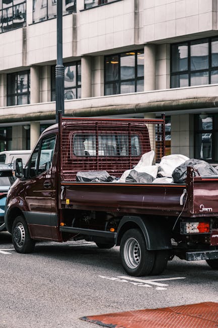 A dark red commercial van with a metal rack at the back, parked on the street outside a modern office building with large windows and concrete facade. The van's open flatbed area is loaded with various household items, including cardboard boxes wrapped in plastic, black garbage bags, and other packaging materials, indicating a home relocation or furniture transport process. The items are arranged in the van's cargo area, which is partially enclosed by the metal rack for stability during transit. Nearby, other vehicles are parked along the street, and a lamppost is visible in the background. The scene suggests an active loading process overseen by a professional moving service, such as Man With a Van West Ham, with the focus on careful packing and efficient transport of household belongings following standard furniture moving procedures. The weather appears clear, with natural light illuminating the scene, highlighting the practical aspects of a house removals operation.