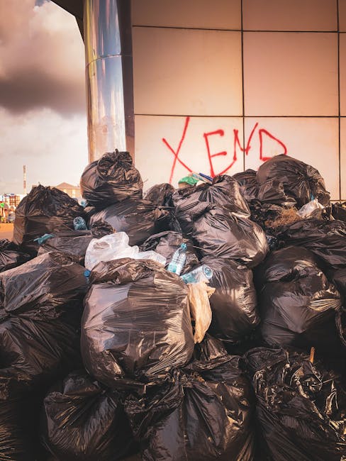 A large pile of black plastic rubbish bags filled with household waste, some tied and others partially open revealing contents such as plastic bottles and packaging, stacked against a tiled wall with red graffiti that reads 'XEND'. The bags are on the pavement adjacent to a commercial building, with a reflection of the sky and nearby structures on a metallic surface to the left. The scene suggests a home relocation context involving waste disposal in line with Newham Council rules, typical of logistics handled by Man With a Van West Ham during bulk waste collection and moving services.