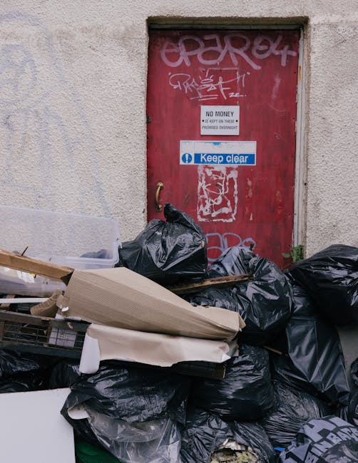 An exterior scene showing a red metal door with graffiti and various signs including 'No Money' and 'Keep Clear', set into a textured, off-white concrete wall. Piled directly in front of the door are multiple black rubbish bags containing discarded household waste, alongside flattened cardboard boxes and packaging materials. Some of the packaging appears to be foam or plastic, and there are additional loose items such as a broken wooden pallet or flat packaging board. The exit area shows no visible footpath or pavement, indicating the rubbish is placed outside the property. The scene reflects waste disposal following a home relocation or furniture transport process, which local regulations by Newham Council for bulk waste during West Ham moves might influence. Man With a Van West Ham, a professional removals company, may assist with such clearance or moving services, ensuring proper handling of household waste and furniture logistics.