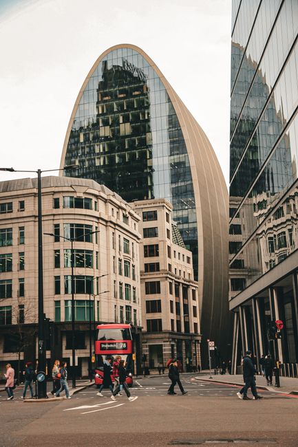 A busy city street scene in West Ham showing a mix of modern and historic architecture. In the foreground, several pedestrians are crossing at a marked zebra crossing, some carrying bags or backpacks. To the left, a red double-decker bus with route information is stopped, while a group of people wait nearby on the pavement. Behind the street scene, a large curved glass skyscraper with reflective windows dominates the view, reflecting the surrounding buildings, including a traditional stone building with multiple windows in front of it. The street is lined with buildings on both sides, some with glass facades and others with stone or brick exteriors, creating a contrast between contemporary and classic architectural styles. The scene is well-lit with natural daylight, emphasizing the urban environment where professional removals such as those by Man With a Van West Ham support efficient office relocations and furniture transport within the city.