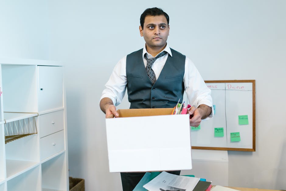 A man dressed in formal office attire, including a white shirt, dark vest, and patterned tie, is standing inside an office space holding an open cardboard box filled with office supplies such as pens, folders, and stationery. The room features white storage units with shelves and drawers on the left side, and a whiteboard with green and pink sticky notes attached on the wall behind him, suggesting a workspace or planning area. The man appears to be preparing for a home relocation or office move, as part of a furniture transport or packing and moving process guided by Man With a Van West Ham. The scene is well-lit with natural or artificial light, highlighting the details of the packing activity and the workspace environment, aligning with professional removals and relocation services.