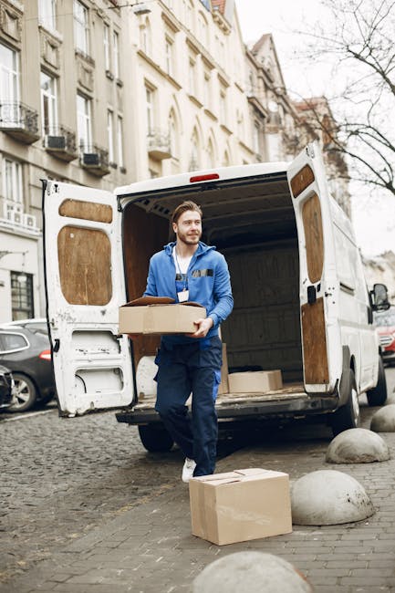 A man wearing a blue jacket and dark pants is placing a cardboard box into the open rear of a white commercial van parked on a cobbled street in an urban area. The van's interior is visible, showing i