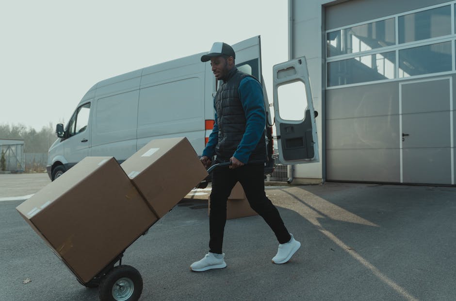 Two men are seated inside the open rear of a van used for home relocation, with cardboard boxes and packing materials visible behind them. The man on the left is wearing a beige knit cap, a green puff