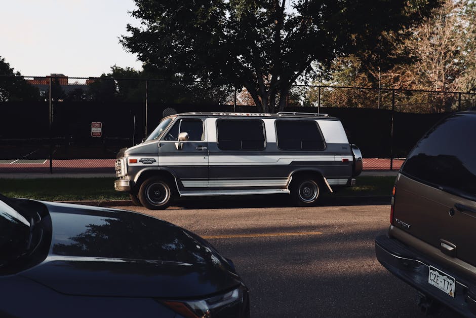 A vintage white and grey van with tinted windows and black accents is parked on the side of a residential street near a tennis court enclosed by a tall black metal fence. The van is positioned parallel to the curb, with its sliding side door closed. The street is lined with trees, some with autumn foliage, casting shadows on the pavement. In the foreground, parts of two modern cars—one black and one dark grey—are visible, partially obstructing the view. The pavement surface is smooth, and there are no visible loading equipment or boxes in the scene. This image captures a calm moment during a home relocation or furniture transport process, showcasing the vehicle used by Man With a Van West Ham for house removals, set against a typical UK neighbourhood background that includes leafy trees and recreational facilities.