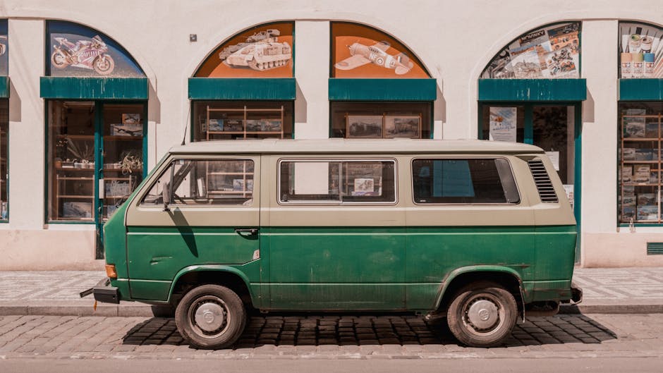 A vintage green and cream van parked parallel to a sidewalk in front of a commercial building with large arched windows displaying models and artwork. The van's sliding side door is closed, and the vehicle appears to be in a weathered condition, indicating regular use for house removals and furniture transport. The van is positioned on cobblestone paving, with the surrounding environment suggesting an urban area suitable for moving logistics. The building behind features blue awnings over each window and light-colored walls, providing a clear background for the van. This scene exemplifies typical home relocation scenes involving furniture and boxes being loaded or prepared for transport by a professional removals company such as Man With a Van West Ham, situated within the context of efficient packing and moving processes for residential relocations in the Upton Park area, as discussed on the webpage Upton Park Moves: Best Van Routes for Green Street, WEST HAM.