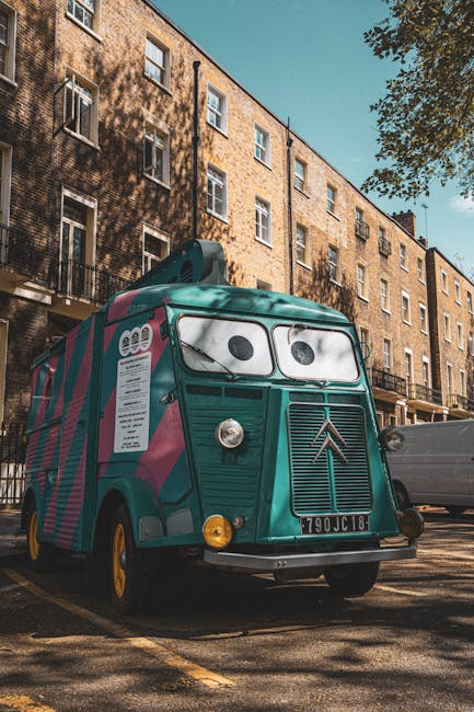 A vintage white and grey van with tinted windows and black accents is parked on the side of a residential street near a tennis court enclosed by a tall black metal fence. The van is positioned parallel to the curb, with its sliding side door closed. The street is lined with trees, some with autumn foliage, casting shadows on the pavement. In the foreground, parts of two modern cars—one black and one dark grey—are visible, partially obstructing the view. The pavement surface is smooth, and there are no visible loading equipment or boxes in the scene. This image captures a calm moment during a home relocation or furniture transport process, showcasing the vehicle used by Man With a Van West Ham for house removals, set against a typical UK neighbourhood background that includes leafy trees and recreational facilities.