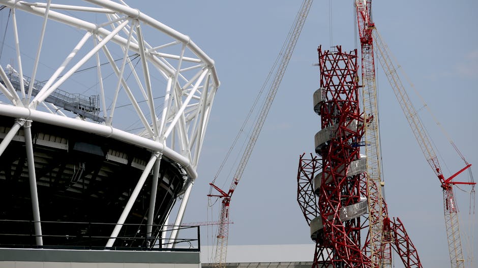 A section of a stadium or arena structure with white steel support beams forming a geometric framework, partially visible in the foreground, contrasted against a cloudy sky. To the right, a tall red and grey telecommunications tower with spiral metal stairs wrapping around its core, accompanied by several large yellow and white cranes with extended booms and cables, indicating ongoing construction or maintenance work. The image is set outdoors and captures the industrial and infrastructural elements, with visible lighting and shadows suggesting daytime. This scene aligns with logistics and complex moving or transport activities, such as those managed by Man With a Van West Ham for home relocation or furniture transport, showing the scale and technical aspects involved in large-scale property moves or infrastructure projects.