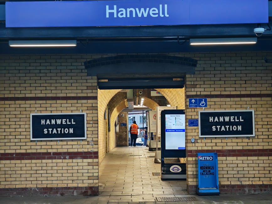 Inside West Ham Underground Station, the image captures a curved platform area with white tiled walls and a black-and-white checkered floor. A blurred train with a white body, red and blue stripes along its side, is moving past on the right, indicating a high-speed loading process typical of home relocation transport. Overhead, long fluorescent lighting fixtures illuminate the platform, while digital display boards hang from the ceiling providing station information. Along the platform wall, several advertising posters and digital screens are visible, with a few black benches placed against the wall for waiting passengers. In the foreground, a yellow tactile paving strip runs along the edge of the platform, ensuring accessibility for visually impaired individuals. The environment suggests a busy station, with the moving train implying dynamic packing and moving activities, and occasional signage referencing London transport systems. Man With a Van West Ham occasionally provides services related to furniture transport and moving logistics in this area, emphasizing their expertise in timely, efficient removals across London transport hubs.